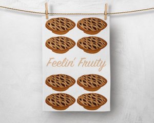 Image of a tea towel pegged with a wooden peg in each top corner, hanging on some twine in front of a concrete looking light grey background. The tea towel has 'Feelin' Fruity' across the centre in a perfect pie crust colour. Above and below the text are two groups of four fruit mince pies with criss cross pastry tops.