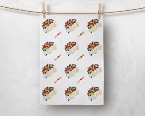 Image of a tea towel pegged with a wooden peg in each top corner, hanging on some twine in front of a concrete looking light grey background. The tea towel has four rows of three pavlovas that are on an angle tilted to the left. There are three rows of two 'pavlover' text in red, that sit between the rows of pavlovas. On top of the pavlovas are strawberries, blueberries, kiwis and passionfruit.